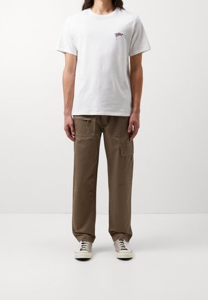 T-shirt en coton blanc avec un petit emblème coloré sur la poitrine, associé à un pantalon cargo loose marron avec poches et cordon de serrage. Baskets légères.