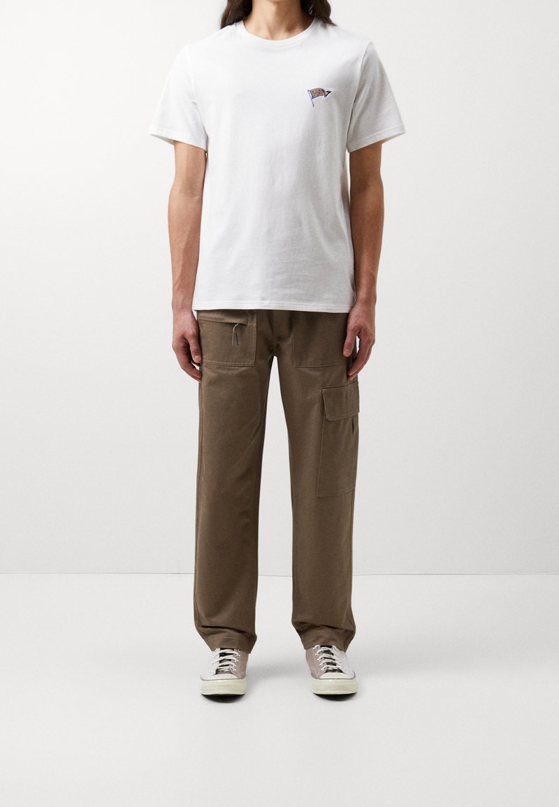 Hvid bomulds t-shirt med lille farverigt emblem på brystet, parret med løse brune cargo-bukser med lommer og snor. Lyse sneakers.