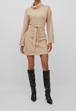 Personne portant une robe-pull beige à manches longues ceinturée avec un col bénitier et des bottes noires montantes à talons, debout sur un fond gris.