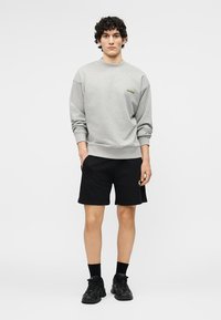 Ung mann står iført en grå hettegenser, svarte shorts, svarte sokker og svarte joggesko mot en ensfarget hvit bakgrunn.