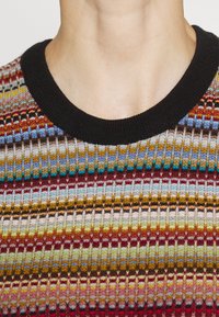 Pull en tricot multicolore présentant des rayures horizontales dans des nuances de rouge, bleu, jaune et gris, avec un col rond côtelé noir.