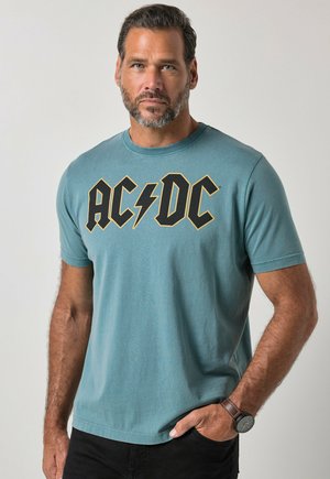 Mężczyzna z brodą w turkusowym t-shircie AC/DC i ciemnych spodniach, stojący z jedną ręką w kieszeni na jednolitym tle.
