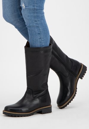Jambes en jean skinny bleu portant de grandes bottes en cuir noir avec fermetures éclair latérales et semelles épaisses à relief sur fond blanc.