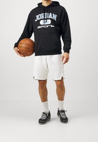 Felpa nera con cappuccio con grafiche "JORDAN" e "SPORT", pantaloni corti in rete bianchi, pallone da basket e scarpe sportive nere con suole testurizzate.