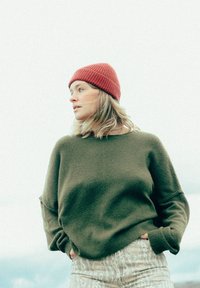 Pull olive vert à manches longues, en tissu texturé ; associé à un pantalon blanc à motifs et un bonnet rouge à côte. Fond neutre.