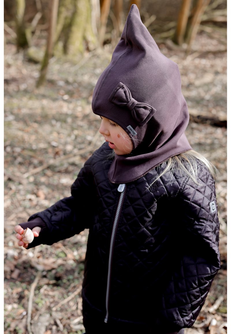 Chaqueta negra acolchada con cremallera plateada, combinada con una capucha ajustada de color púrpura oscuro con detalle de lazo. Niño sosteniendo un pequeño objeto al aire libre.