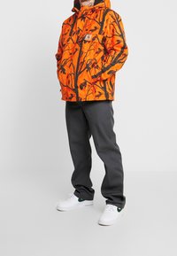 Giacca impermeabile arancione brillante con motivo di alberi e uccelli neri, dotata di tasca frontale. Abbinata a pantaloni grigio scuro e sneaker bianche.