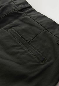 Sötétzöld, tartós anyagú nadrág, sima textúrával, hátsó zsebbel és varrott részletezéssel az illesztések mentén.