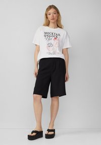 T-shirt graphique blanche avec le texte "MOCKTAIL O'CLOCK" et une illustration de cocktail ; associé à un short noir jusqu'aux genoux et des sandales à plateforme noires.