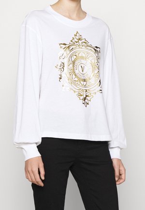 Hvid langærmet skjorte med gyldent, udsmykket Versace Jeans Couture-logo centreret foran, båret med sorte bukser.