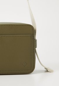 Olive green leather bag with a rectangular shape, rounded edges, cream strap, and subtle logo detail. Smooth texture and minimalistic design.
