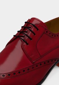 Ένα κόκκινο δερμάτινο παπούτσι με γυαλιστερό φινίρισμα, διαθέτει σχεδιασμό με κορδόνια, διάτρητο μοτίβο κατά μήκος των άκρων και λεία υφή.