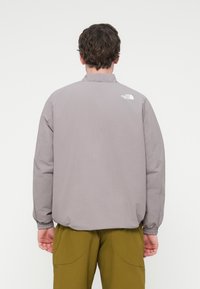 Γκρι, ελαφρύ μπουφάν με ελαστικά μανσέτες και γιακά όρθιο. Φέρει το λογότυπο «The North Face» σε λευκό στο επάνω μέρος της πλάτης.