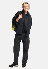Dunkelblaues Fleecebeste mit hohem Kragen, über einem schwarzen Shirt und schwarzen Cargo-Hosen getragen; hält einen gelben Mesh-Rucksack, schwarze Sneakers.