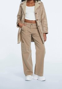 Beige nepbontjas met grote knopen, gedragen over een witte crop top en beige cargobroek, compleet met chunky witte sneakers.