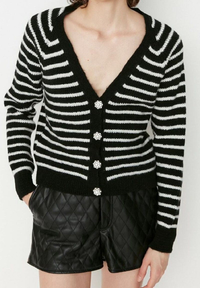 Schwarz-weißer gestreifter Cardigan mit V-Ausschnitt, ausgestattet mit fünf dekorativen Knöpfen. Getragen über gepolsterten schwarzen Shorts.