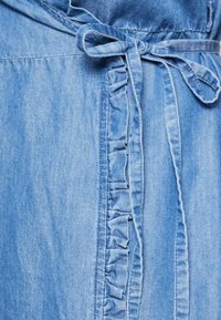 Ljust blå denimplagg med volangkant fram och ett knutet bälte vid midjan, som visar en slät textur och en avslappnad design.