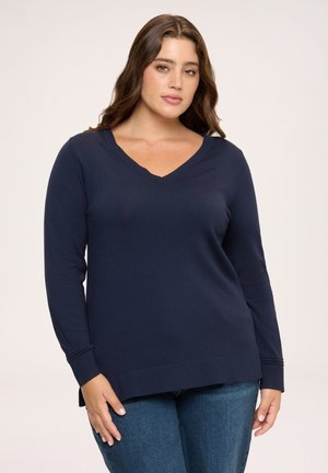 Frau trägt einen marineblauen Pullover mit V-Ausschnitt und langen Ärmeln sowie blaue Jeans, steht vor einem schlichten hellen Hintergrund.