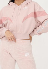 Ljusrosa zip-up hoodie med tonade rosa paneler och vita logodetaljer, kombinerad med matchande ljusrosa byxor med en subtilt mönster.