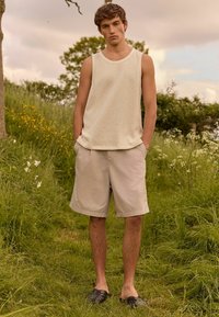 Lys beige strikket tanktop med et struktureret mønster; parret med lyse khakishorts og sorte vævede slip-on sko. Model står på græs.