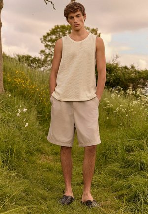 Svetle béžový pletený top bez rukávů s texturovaným vzorem; kombinovaný se světle khaki šortkami a černými tkanými slip-on botami. Modelka stojí na trávě.