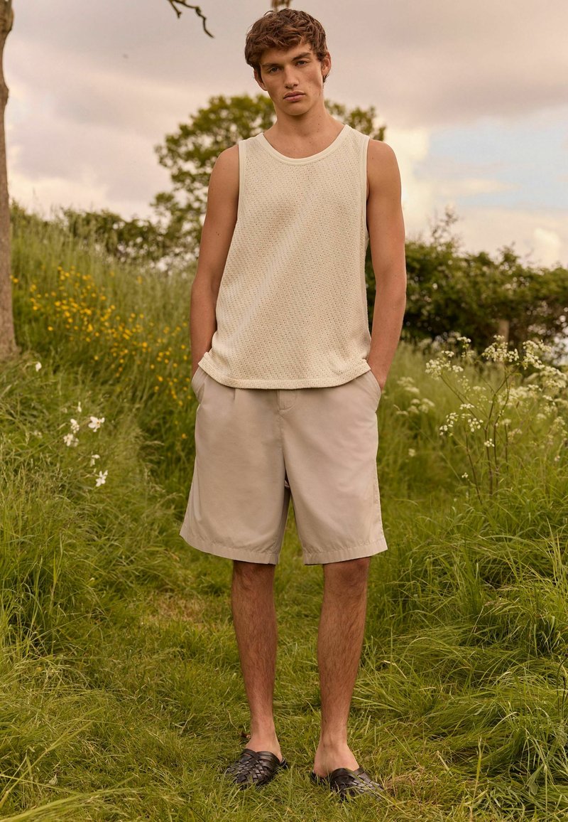 Lys beige strikket tanktop med et struktureret mønster; parret med lyse khakishorts og sorte vævede slip-on sko. Model står på græs.