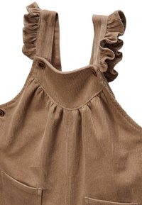 Salopette en velours côtelé marron avec des bretelles à volants, deux poches avant et des boutons en bois pour la fermeture. Matériau texturé et doux.