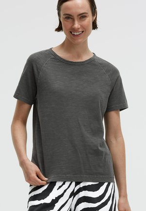 Woman wearing a charcoal gray short-sleeve t-shirt and black and white zebra-striped pants, smiling with hand holding shirt hem.