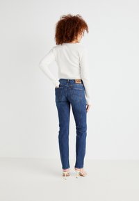 Moteris su garbanotais plaukais, vilkinti baltą ilgomis rankovėmis marškinių viršūnę, Mėlynas Lee džinsus ir baltus aukštakulnius sandalus, stovinti prieš baltą foną.