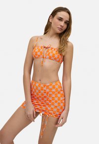 Bikini de un brillante color naranja con un patrón floral, que incorpora un top con lazo frontal y una falda fruncida con ataduras laterales, confeccionado en un tejido ligero y elástico.