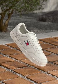 Baskets en cuir blanc avec bout rond, lacets plats, patch logo sur le côté et design minimaliste, posées sur une surface en pierre texturée.
