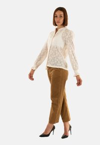 Blusa di pizzo color crema con maniche lunghe e colletto, abbinata a pantaloni in velluto a coste marroni. Tacchi neri completano l'outfit.