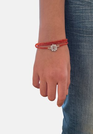 Poignet avec bracelet rouge à trois brins de perles, ornée d'un charme circulaire en argent, placé à côté d'une personne portant un jean bleu.