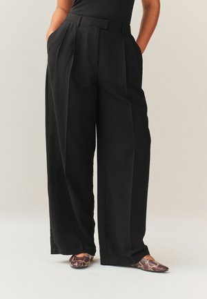 Personne portant un pantalon noir à jambes larges avec des poches latérales et des chaussures plates pointues à motif peau de serpent, debout devant un fond uni.