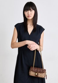 Brown and tan quilted handbag with a chain strap, featuring a prominent clasp. Worn by a model in a dark, v-neck dress.