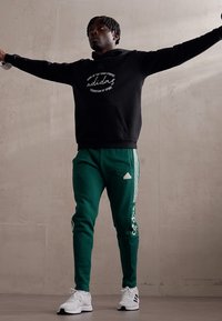 Felpa nera con logo bianco, pantaloni della tuta verdi con strisce bianche e logo, e scarpe da ginnastica bianche con dettagli neri. Il modello sta in piedi con le braccia alzate.