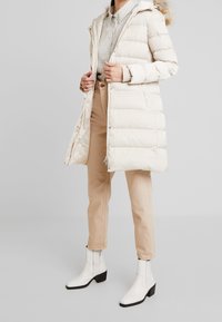 Beige puffer coat with a hood and faux fur trim, featuring quilted design and metallic snaps, paired with light brown pants and white ankle boots.