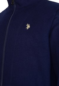 Námornícka zipsová bunda vyrobená z hladkej tkaniny. Má vysoký golier, centrálny zips a malé zlaté logo na ľavom hrudníku.