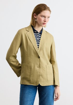 Kobieta ubrana w beżową marynarkę na jeden guzik, granatową koszulkę polo w paski oraz niebieskie dżinsy, stojąca z jedną ręką za plecami.