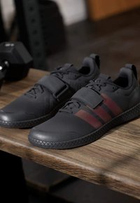 Para czarnych butów sportowych z bordowymi paskami i rzepami leżących na drewnianej ławce obok hantle w siłowni.