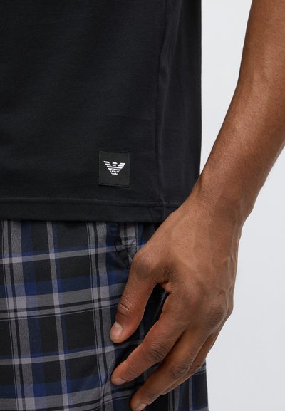 Μαύρο βαμβακερό μπλουζάκι με ορθογώνιο μοτίβο λογότυπου που απεικονίζει ένα στυλιζαρισμένο αετό. Φοριέται με μπλε και γκρι καρό παντελόνι.