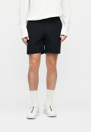 Muž oblečený v čiernych šortkách, bielej dlhoruká košeľa, bielych ponožkách a bielych teniskách s farebnými akcentmi loga stojaci na bielom pozadí.