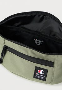 Πράσινη υφασμάτινη θήκη με μαύρο εσωτερικό με φερμουάρ, διαθέτει μια μικρή εξωτερική τσέπη και ένα αυτοκόλλητο λογότυπο Champion. Μοντέρνο μέγεθος, ανθεκτική υφή.