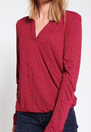 Kvinde iført en rød langærmet bluse med lille hvidt prikkemønster og mørkeblå jeans foran en ensfarvet hvid baggrund.