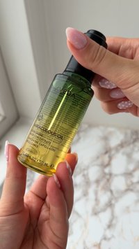 Mani con unghie curate con smalto rosa e bianco tengono una bottiglia verde e gialla di Aveda Miraculous Oil concentrato per capelli.