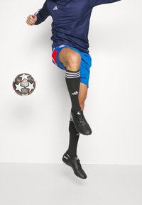 Mužský fotbalista v námořnickém topu, modrých šortkách a černých kopačkách Adidas skáče, aby ovládl černobílý fotbalový míč s hvězdicovým vzorem.