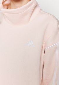 Sweat-shirt rose clair en tissu texturé avec un col montant. Présente un logo Adidas blanc et des bandes accentuées blanches sur les manches.