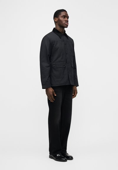 Μαύρο βαμβακερό μπουφάν με κούμπωμα με κουμπιά, δύο εμπρόσθιες τσέπες και υφασμένη γιακά, συνδυασμένο με μαύρο παντελόνι φαρδιάς γραμμής και μαύρα παπούτσια.