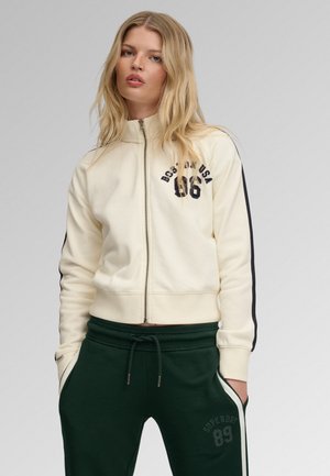 Blond vrouw die een crème zip-up jack draagt met zwarte strepen en donkergroene joggingbroek met witte strepen en zakken.