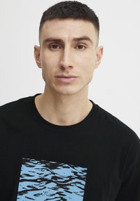 Juoda marškinėliai su mėlynos vandens bangavimosi grafika stačiakampio formos. Lygi medžiaga, apvaliu kaklu ir trumpomis rankovėmis.